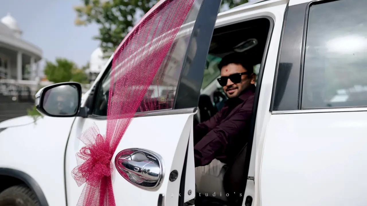 The groom, in sunglasses, steps out of a white car tied with a magenta ribbon